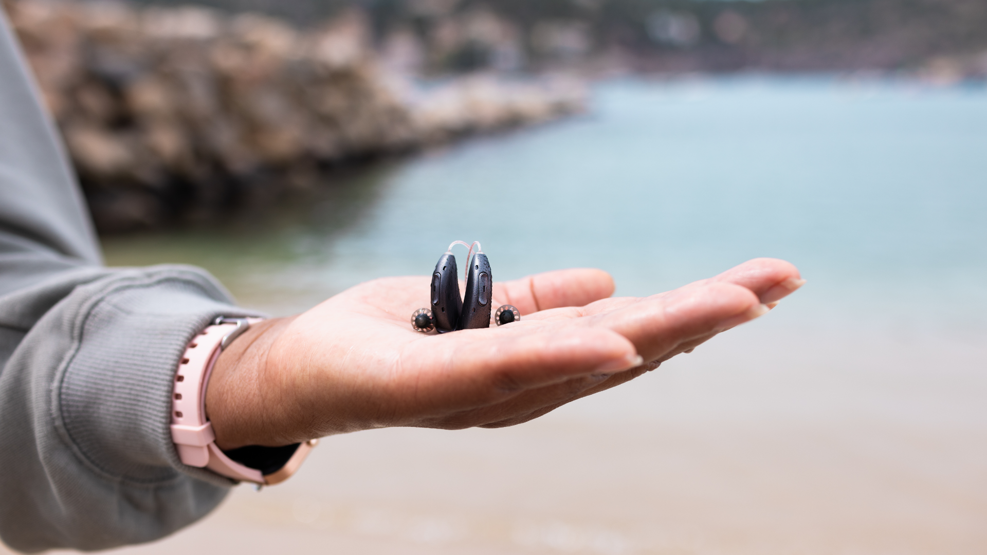 Mão segurando um par de aparelhos auditivos discretos, com o mar ao fundo